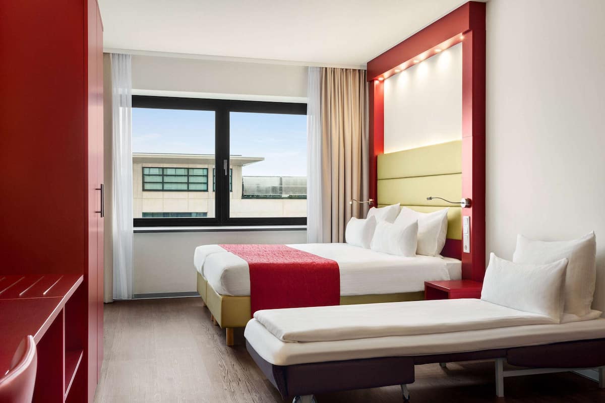 Une chambre d’hôtel avec deux lits, un banc rouge, une fenêtre donnant sur un immeuble et un mur rouge.