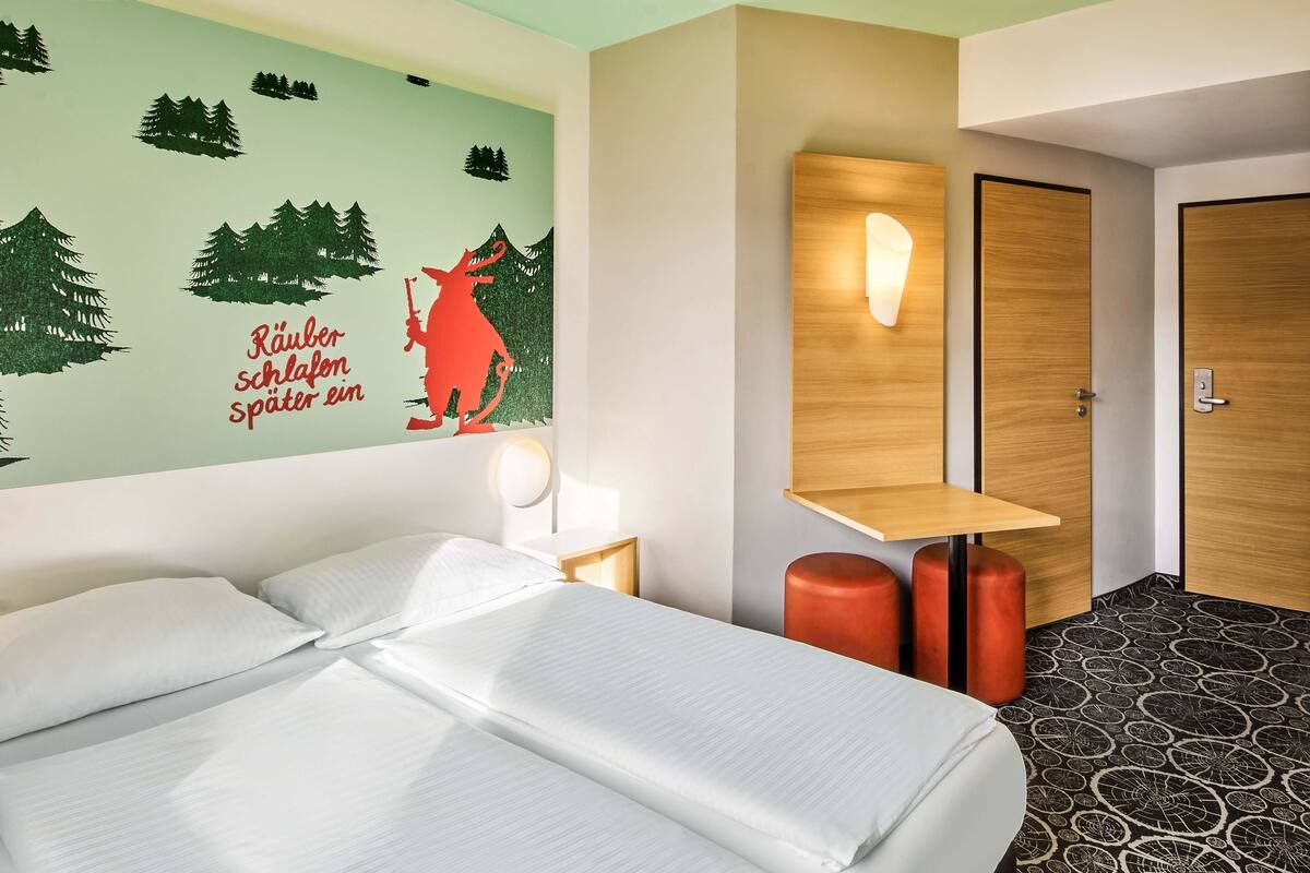 A hotel room with a bed, a wall mural, a bedside table with a lamp, a wooden shelf, and a door.