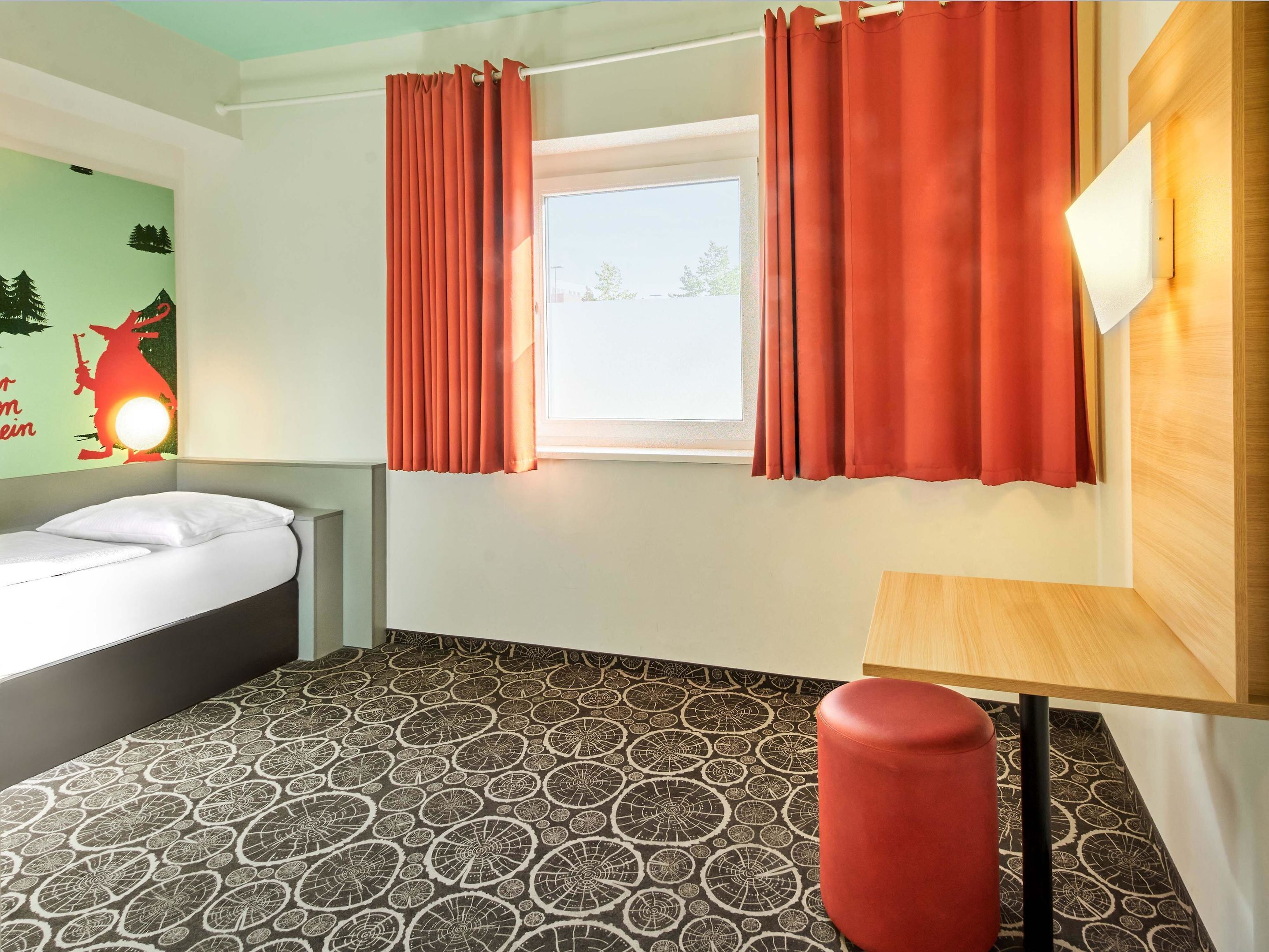 Ein Hotelzimmer mit einem Bett, einem Fenster mit Vorhängen, einem gemusterten Boden und einem kleinen Tisch.