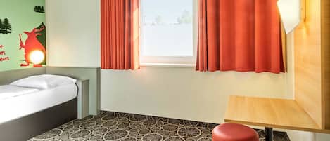 Una habitación de hotel con cama, ventana con cortinas, piso estampado y una mesita.
