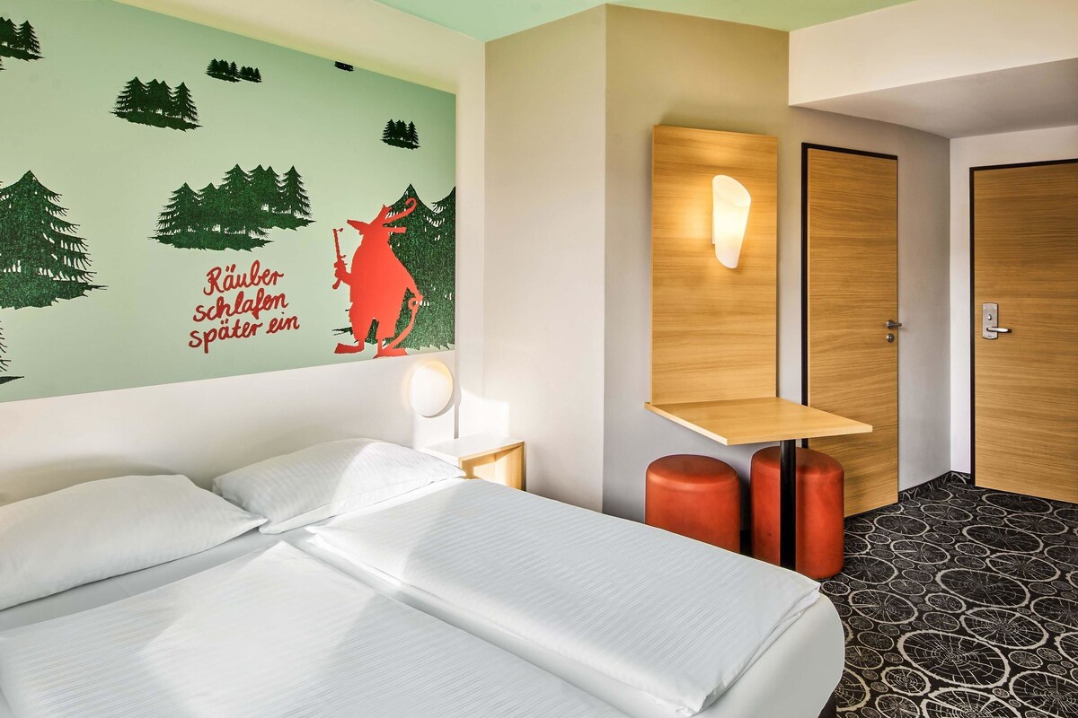 Habitación de hotel con una cama, un mural en la pared, una mesita de noche con lámpara, un estante de madera y una puerta.