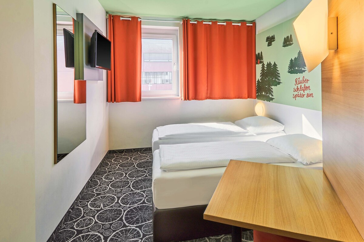 Habitación de hotel con cama, una mesita, televisor montado en la pared y ventana con cortinas.