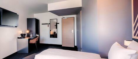 A modern hotel room with a large bed, a flat-screen TV mounted on the wall, a small seating area with a chair and table, and a mirror reflecting the room.