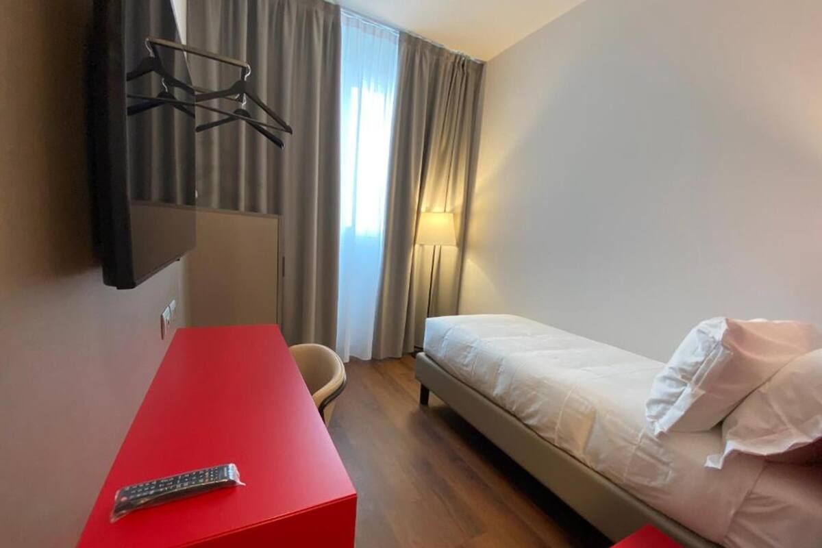 Une chambre d’hôtel avec un lit simple, un bureau rouge, un téléviseur fixé au mur et une fenêtre avec des rideaux.