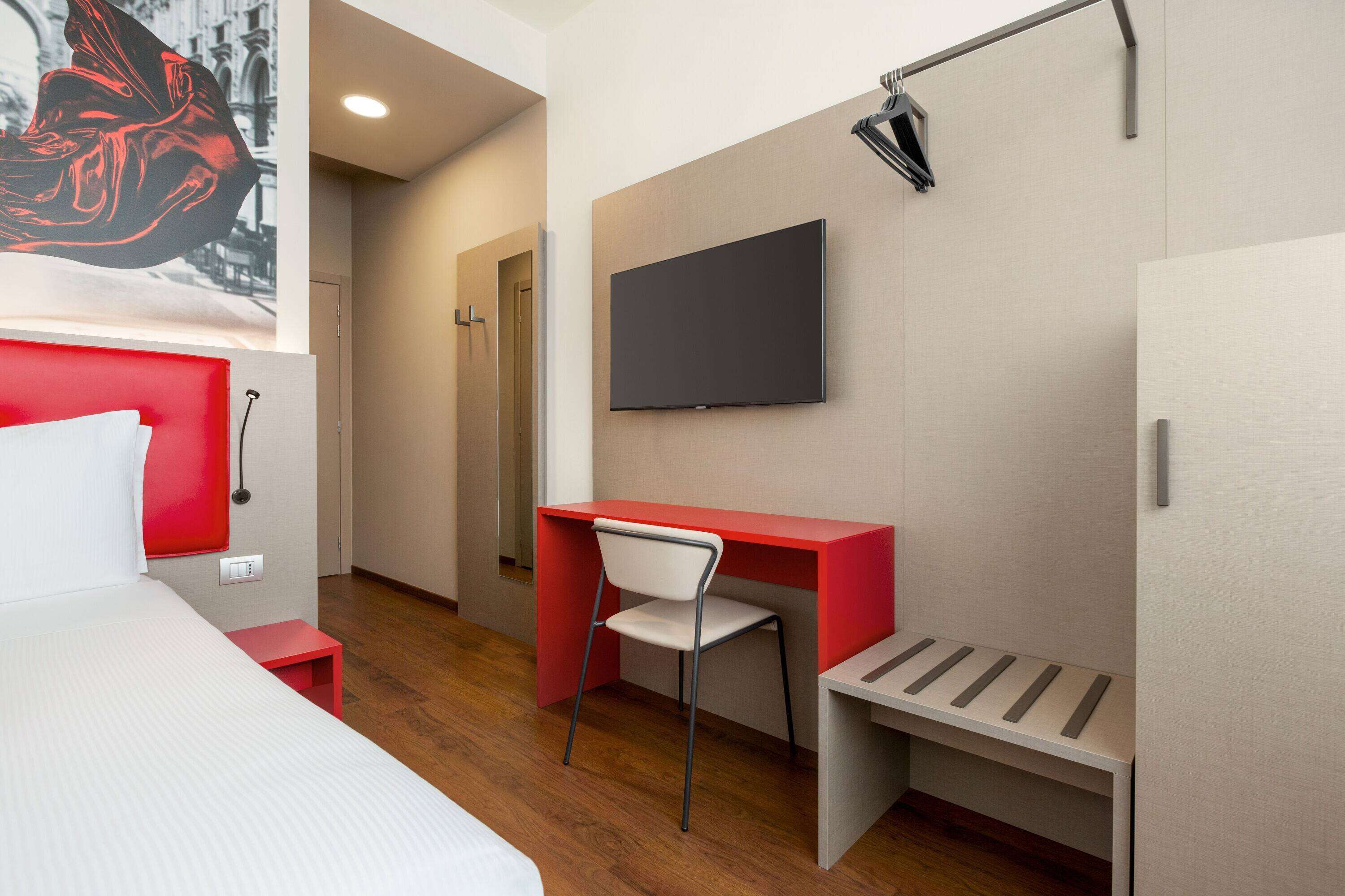 A hotel room with a bed, a desk, a chair, a flat-screen TV, and a painting on the wall.