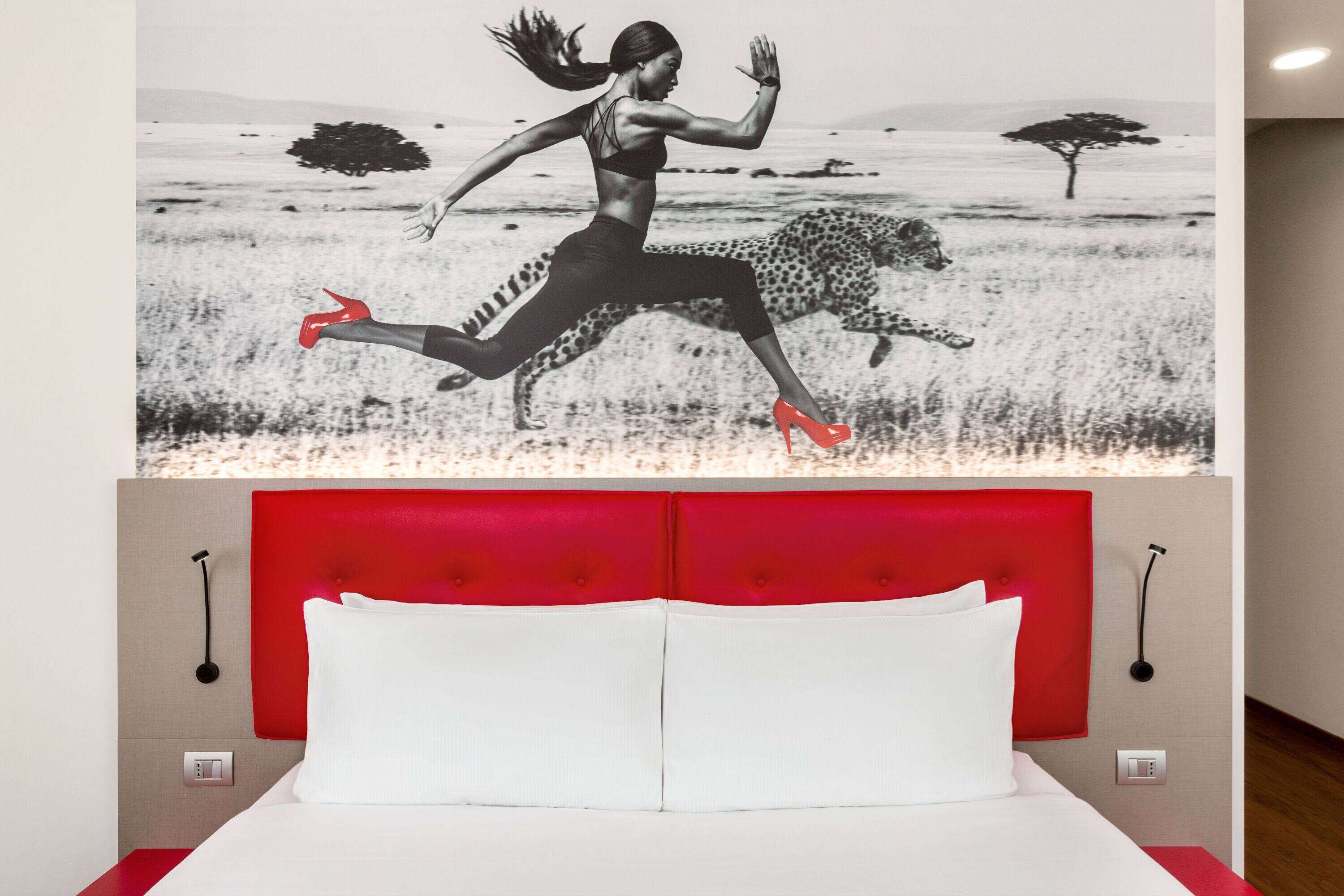 A bedroom with a red headboard, white bedding, and a large mural of a woman running with a leopard.