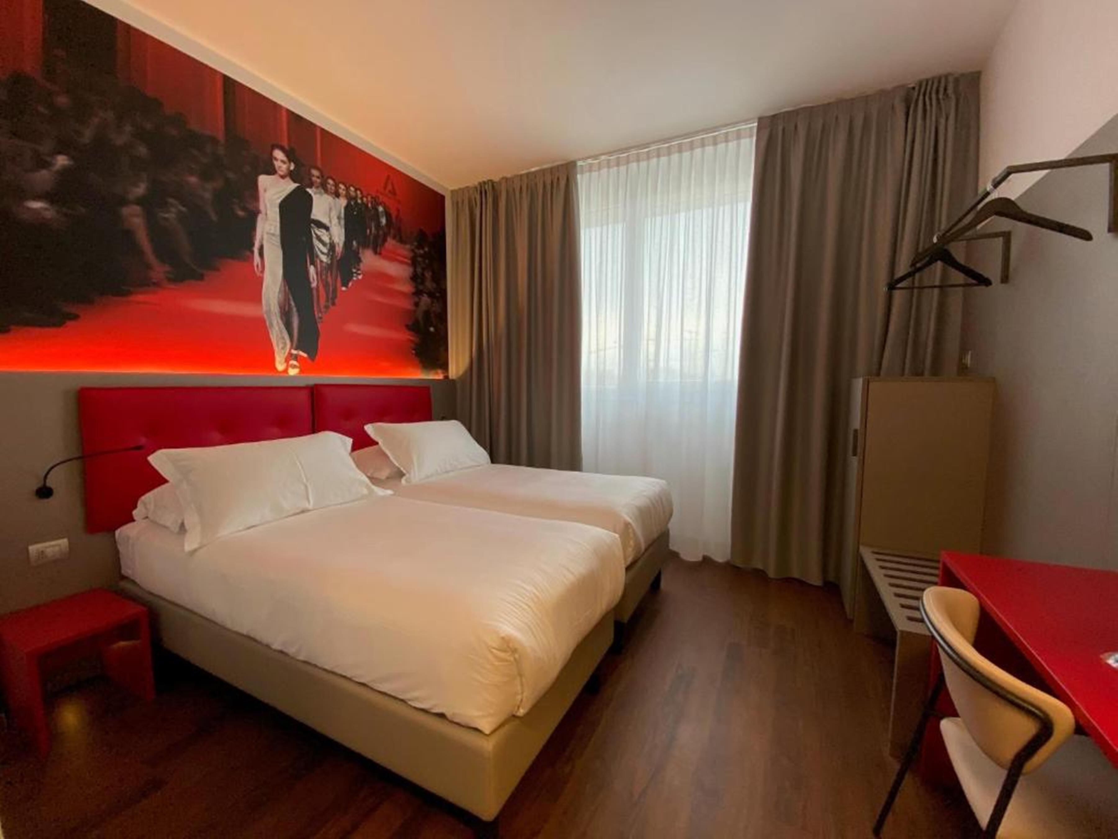 Una habitación de hotel con dos camas, cabecera y escritorio rojos, una silla, un armario y un cuadro grande en la pared.