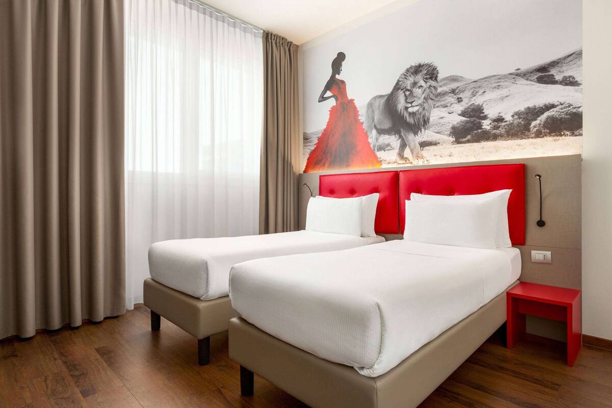 Une chambre d’hôtel avec deux lits, une tête de lit rouge, une petite table de chevet rouge et une grande fresque murale.