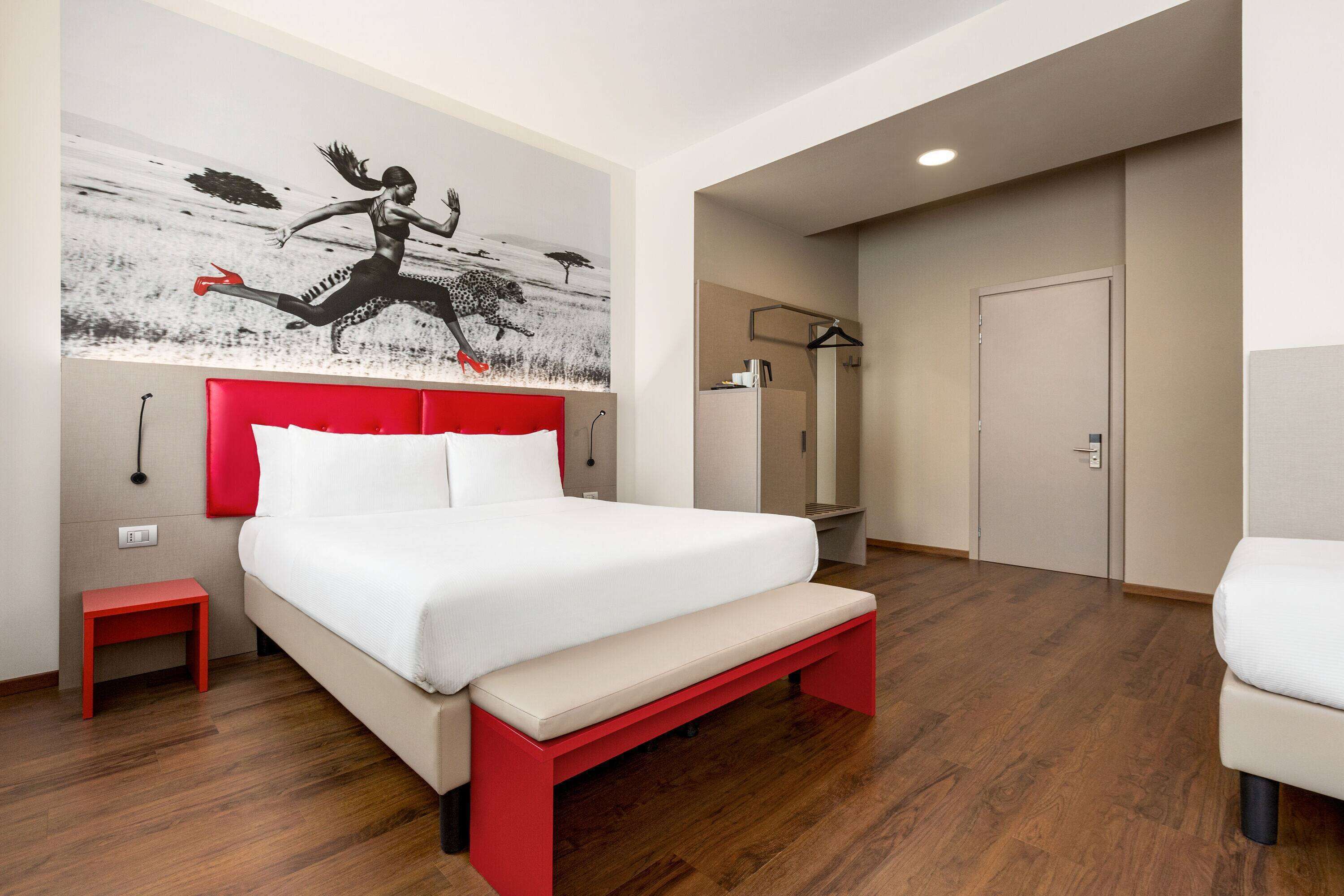 A modern bedroom with a large bed, a red bench, a small red table, a wardrobe, and a mirror.