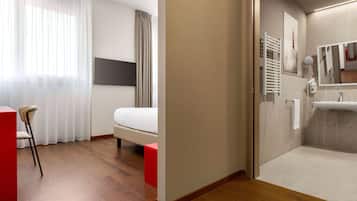 Una camera d'albergo con un letto, una sedia, una scrivania, un bagno con lavandino e specchio, e uno spazio doccia.