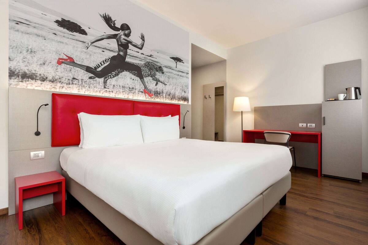 Une chambre d’hôtel avec un grand lit, une table de chevet rouge, un bureau et une œuvre d’art accrochée au mur.