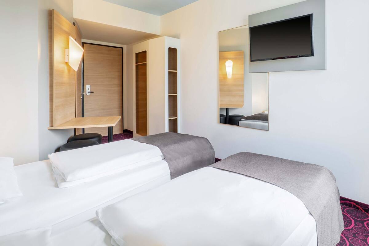 A hotel room with two beds, a TV mounted on the wall, a wardrobe, and a desk with a lamp.