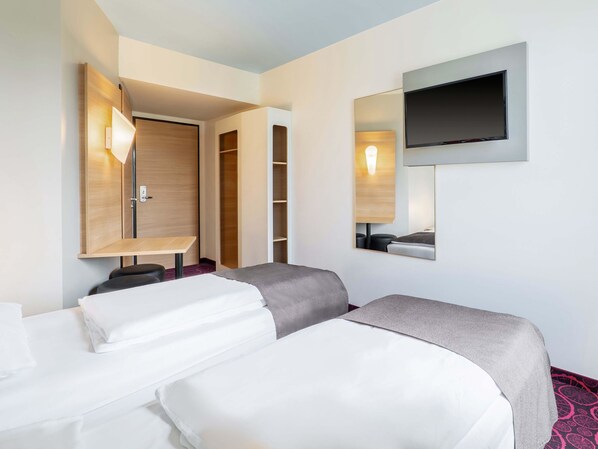 Quarto de hotel com duas camas, uma televisão presa na parede, um guarda-roupa e uma mesa com abajur.