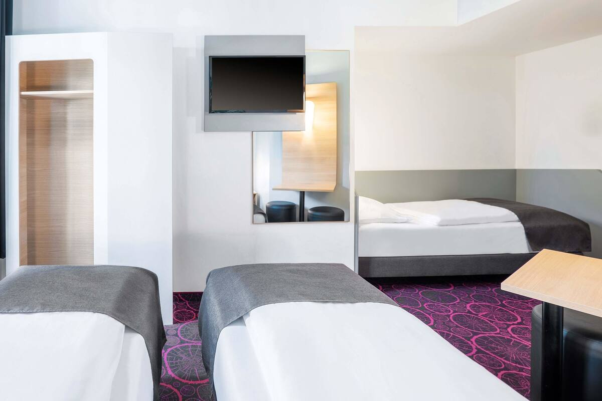 A hotel room with two single beds, a TV mounted on the wall, a bedside table, and a wardrobe.