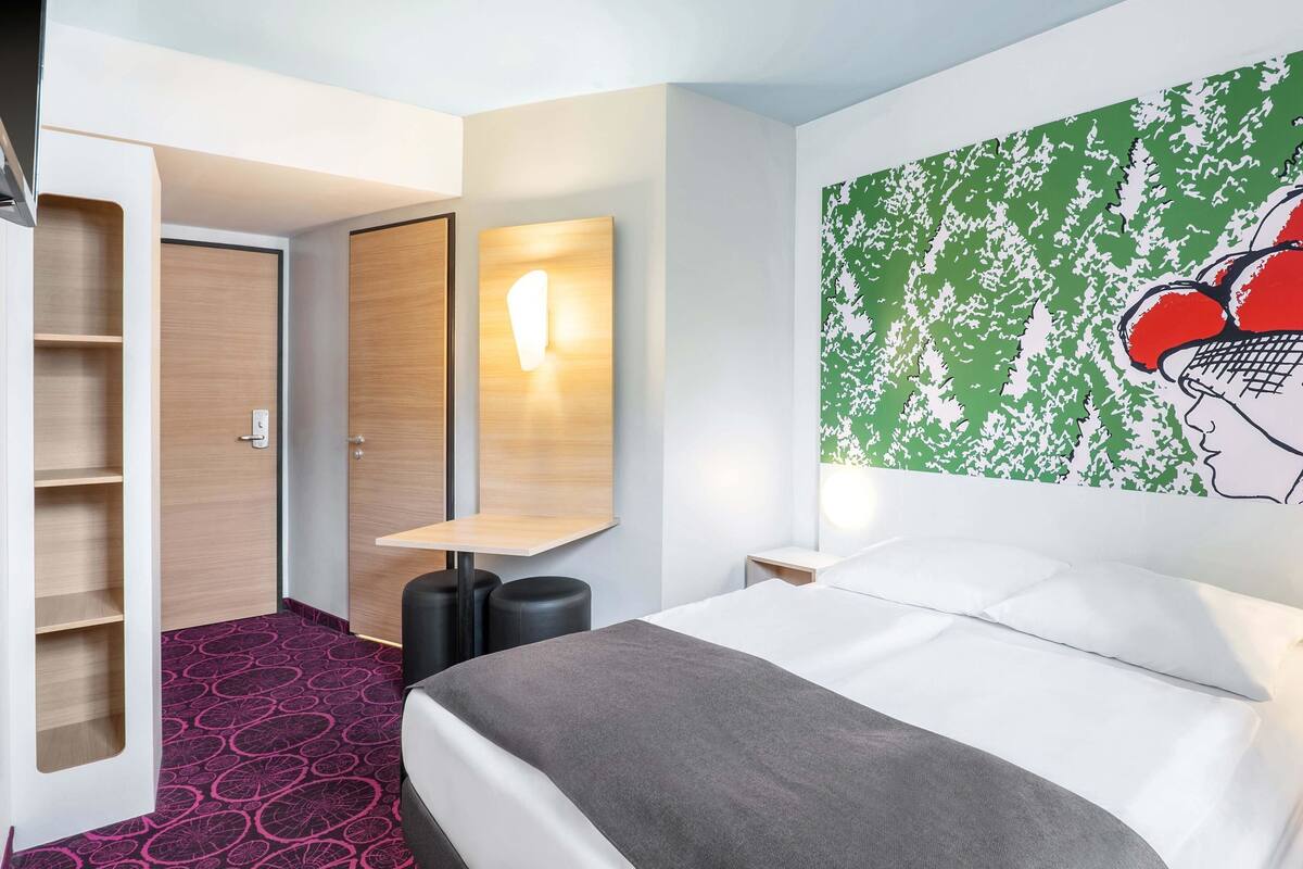 A hotel room with a bed, a wardrobe, a mirror, and a wall with a green and white abstract design.