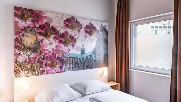 A hotel room with a bed, white bedding, a wall-mounted artwork of pink flowers and a building, and a window with curtains. - B&B Hotel Hildesheim (Hildesheim)
