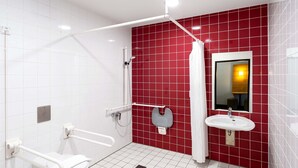 A modern bathroom with white and red tiles, a white toilet, a shower with a white curtain, and a sink with a mirror. - B&B Hotel Hildesheim (Hildesheim)