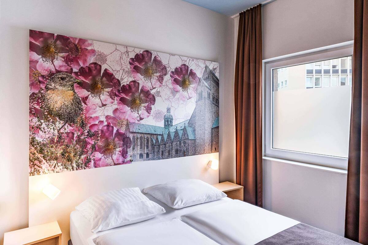 Ein Hotelzimmer mit einem Bett, weißer Bettwäsche, einem Wandbild mit rosa Blumen und einem Gebäude sowie einem Fenster mit Vorhängen.