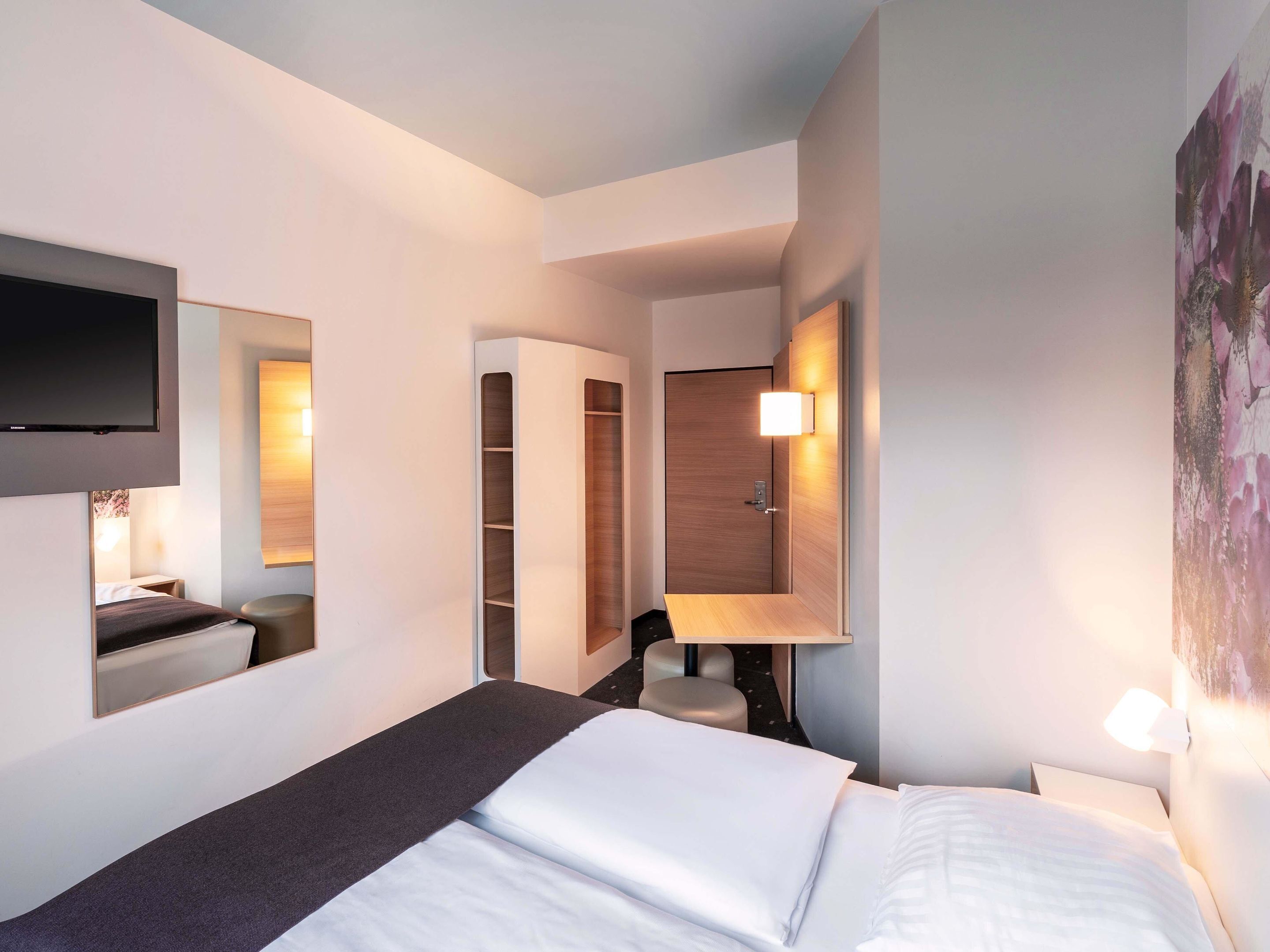 Una habitación de hotel moderna con una cama, un televisor montado en la pared, un armario y un cuadro.