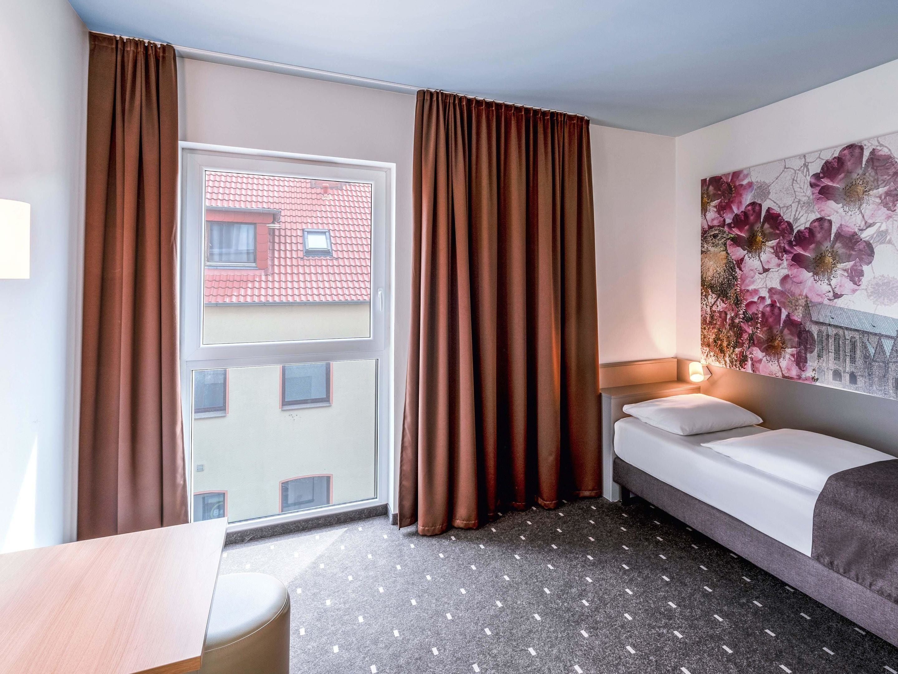 Une chambre d’hôtel avec un lit, un bureau, une fenêtre avec des rideaux et une décoration murale à motifs floraux.