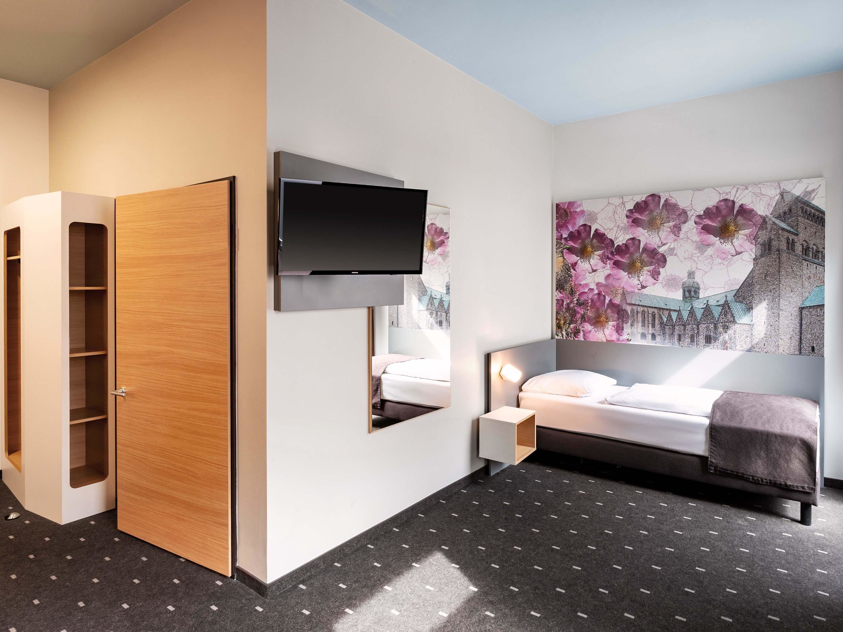 A modern hotel room with a bed, a TV mounted on the wall, a wooden wardrobe, and a large floral artwork on the wall.