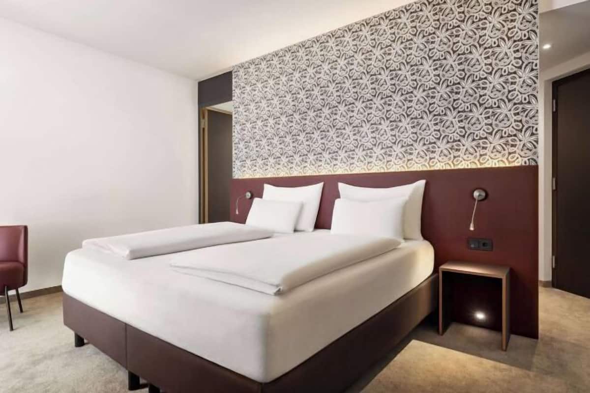 A hotel room with a large bed, a small bedside table, a chair, and a patterned wall.
