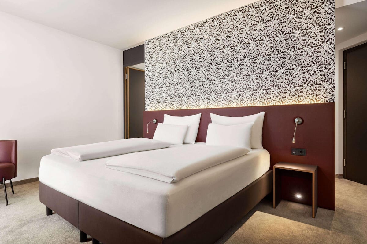 A modern hotel room with a large bed, a small bedside table, a chair, and a decorative wall panel.