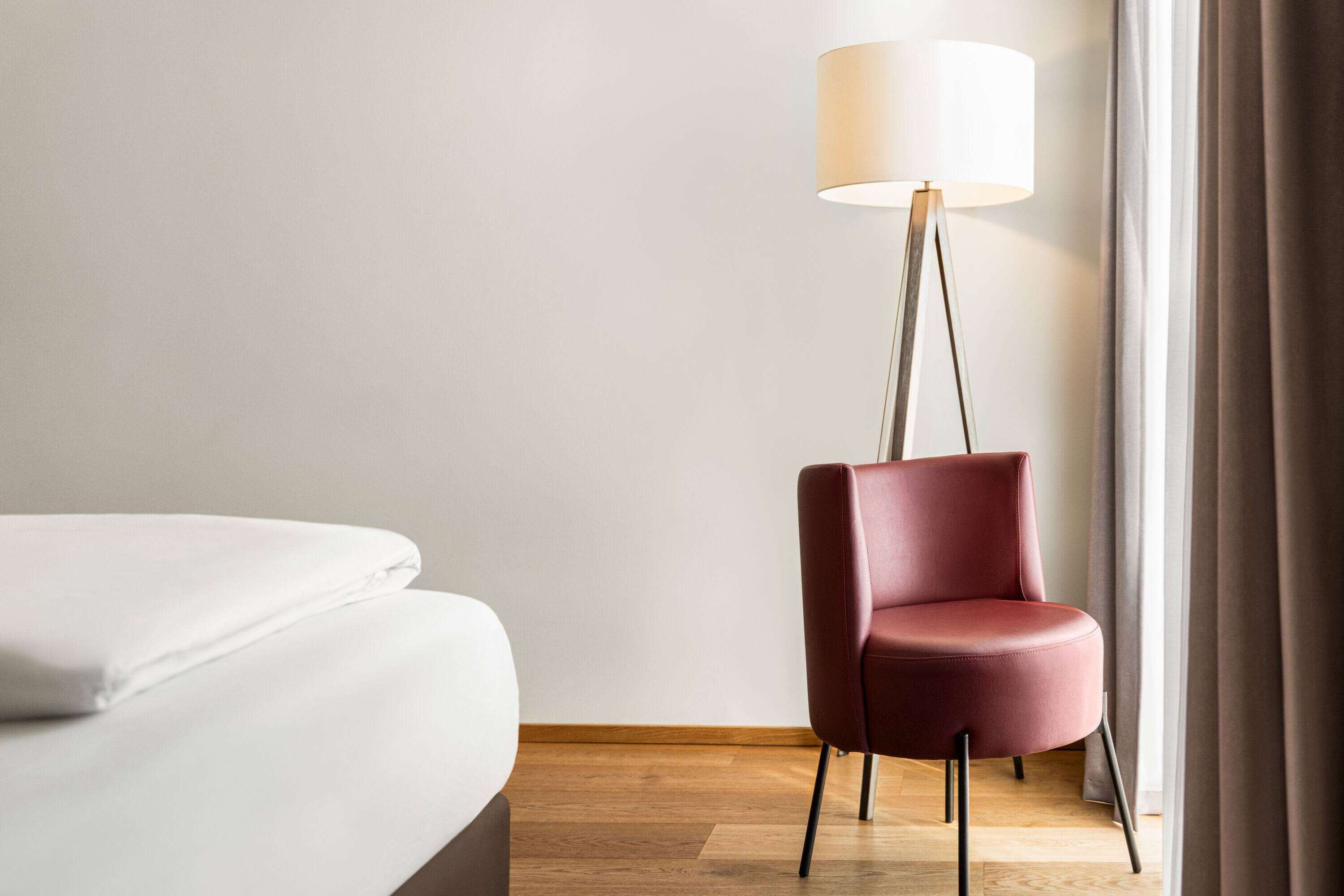 A room with a bed, a red chair, a floor lamp, and a window with curtains.