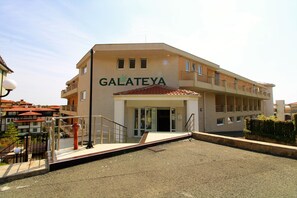 Um edifício com o nome "GALATEYA" na fachada, com teto plano, várias varandas e entrada com portão.