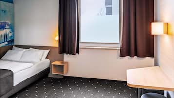 Habitació d'hotel amb un llit, una taula petita, una cadira, una finestra amb cortines i un quadre a la paret.