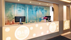 Un comptoir d’accueil d’hôtel orné d’une fresque représentant un paysage urbain, avec deux écrans, un vase de fleurs et le logo de la chaîne B&B Hotels.