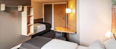 A compact hotel room with a bed, a small desk, a chair, and a wall-mounted shelf.