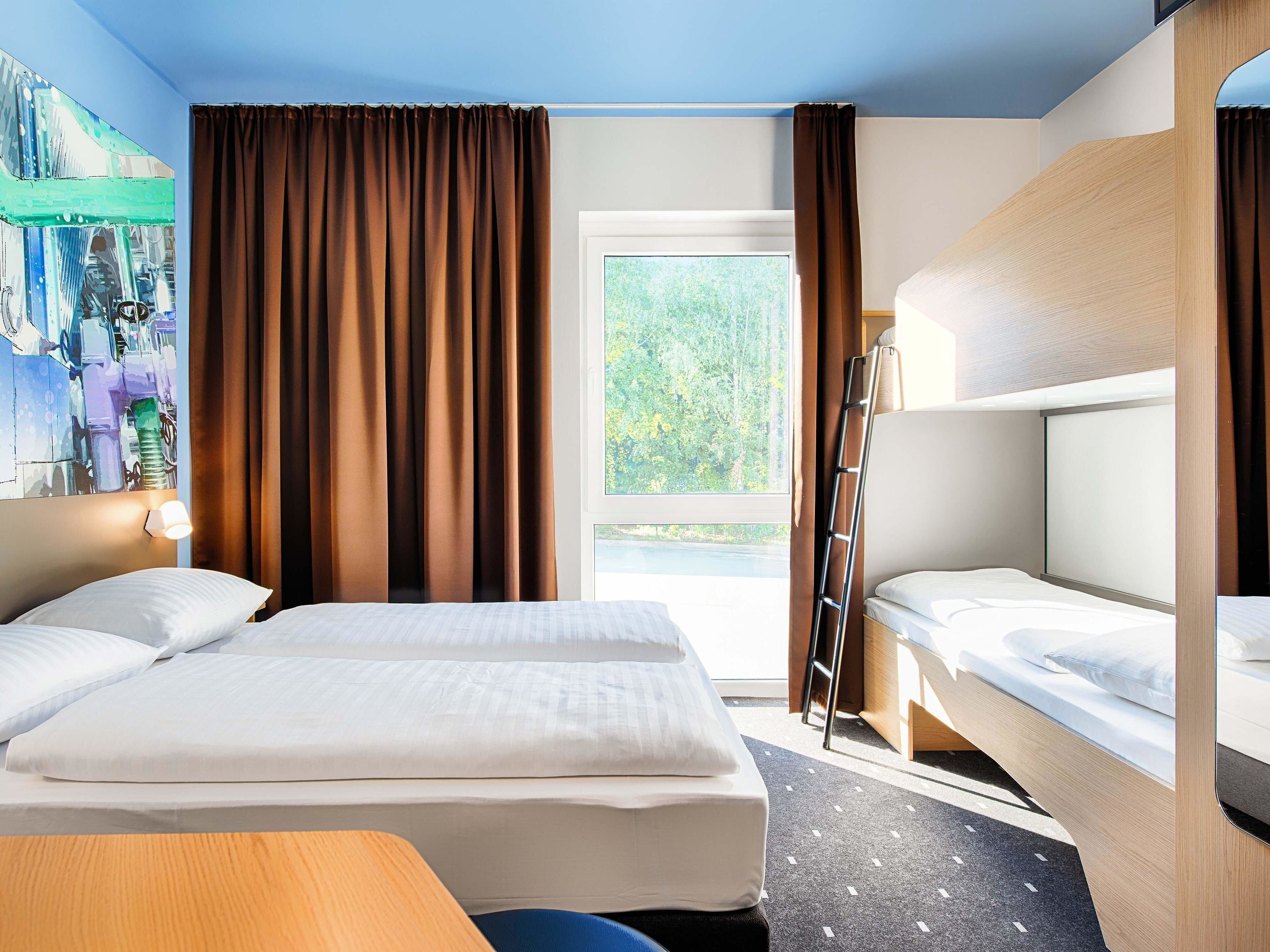 Ein Hotelzimmer mit einem großen Bett, einem Einzelbett, einem Schreibtisch und einem Fenster mit Vorhängen.