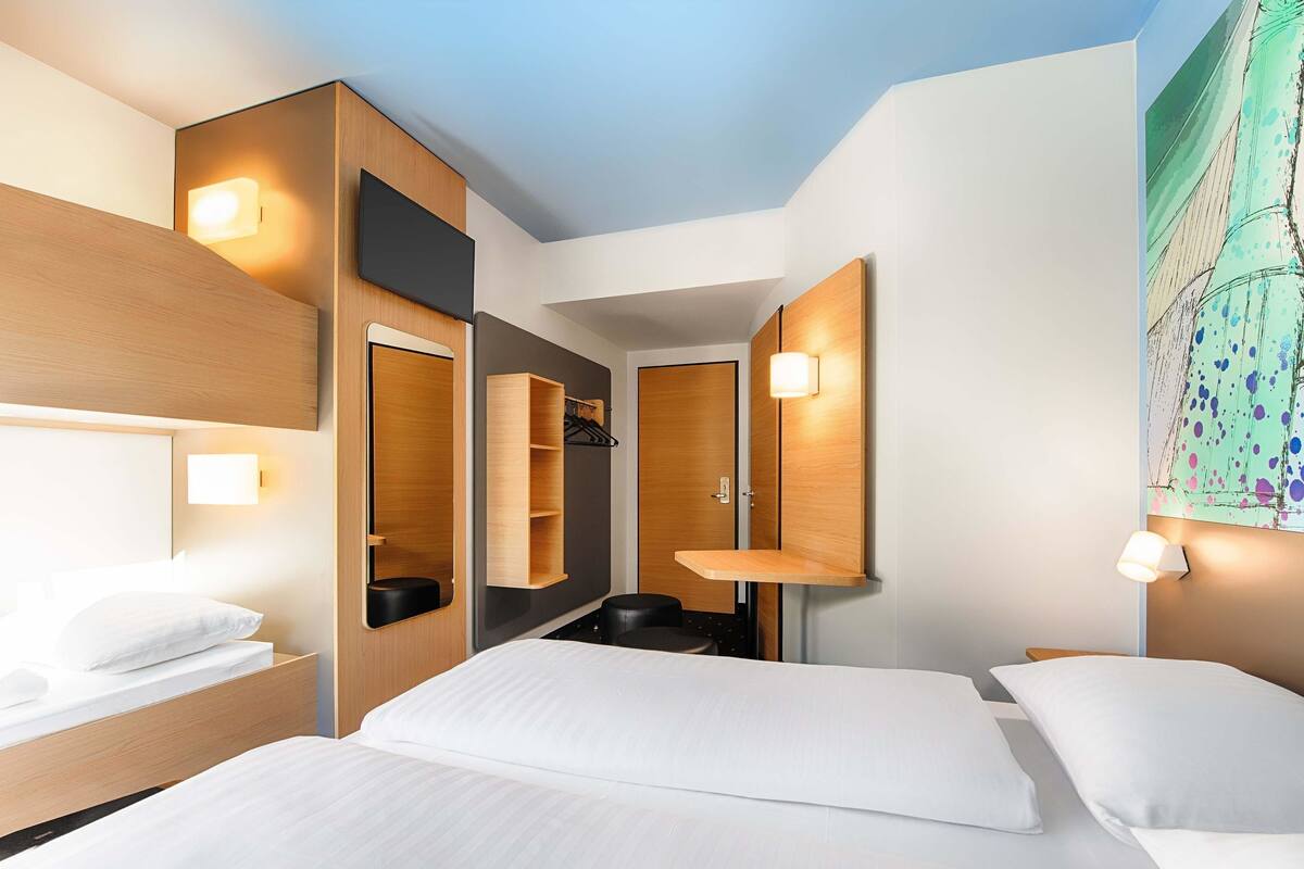 Una habitación de hotel moderna con una cama grande, una mesita de noche, un espejo, un armario y una pequeña zona de estar.