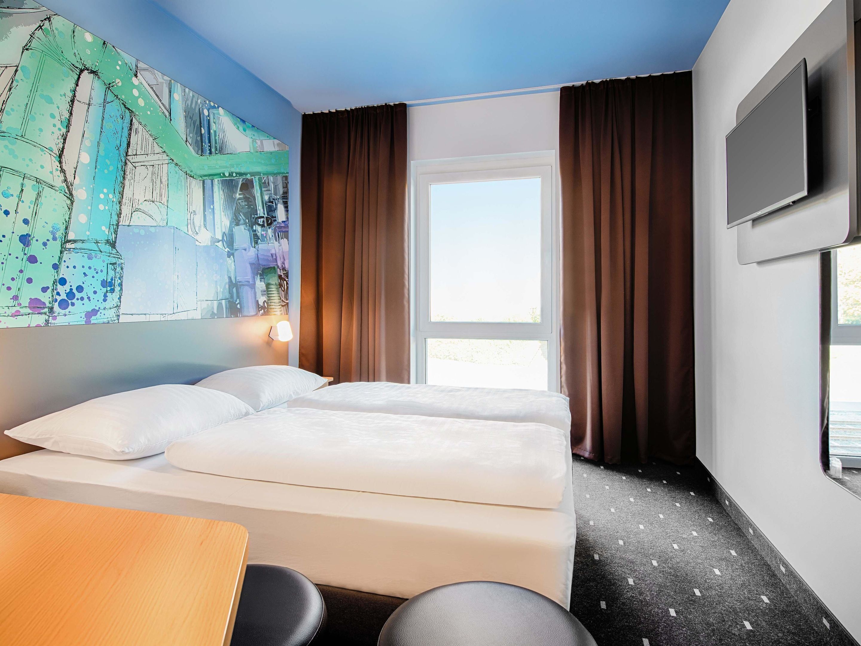 Una camera d'albergo con un letto, una scrivania, una sedia, un quadro appeso al muro e una TV a parete.