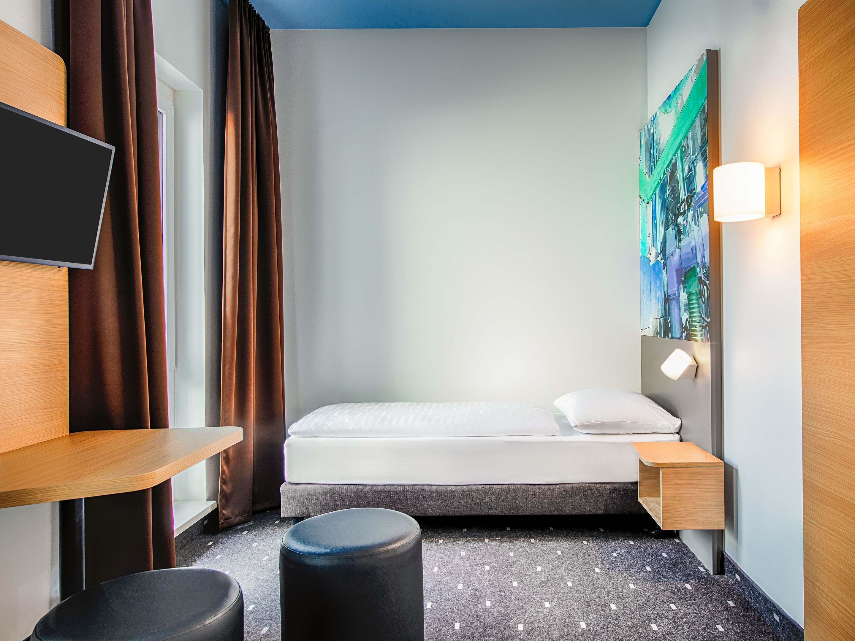 Quarto de hotel compacto com cama, escrivaninha, cadeira, televisão e um quadro na parede.