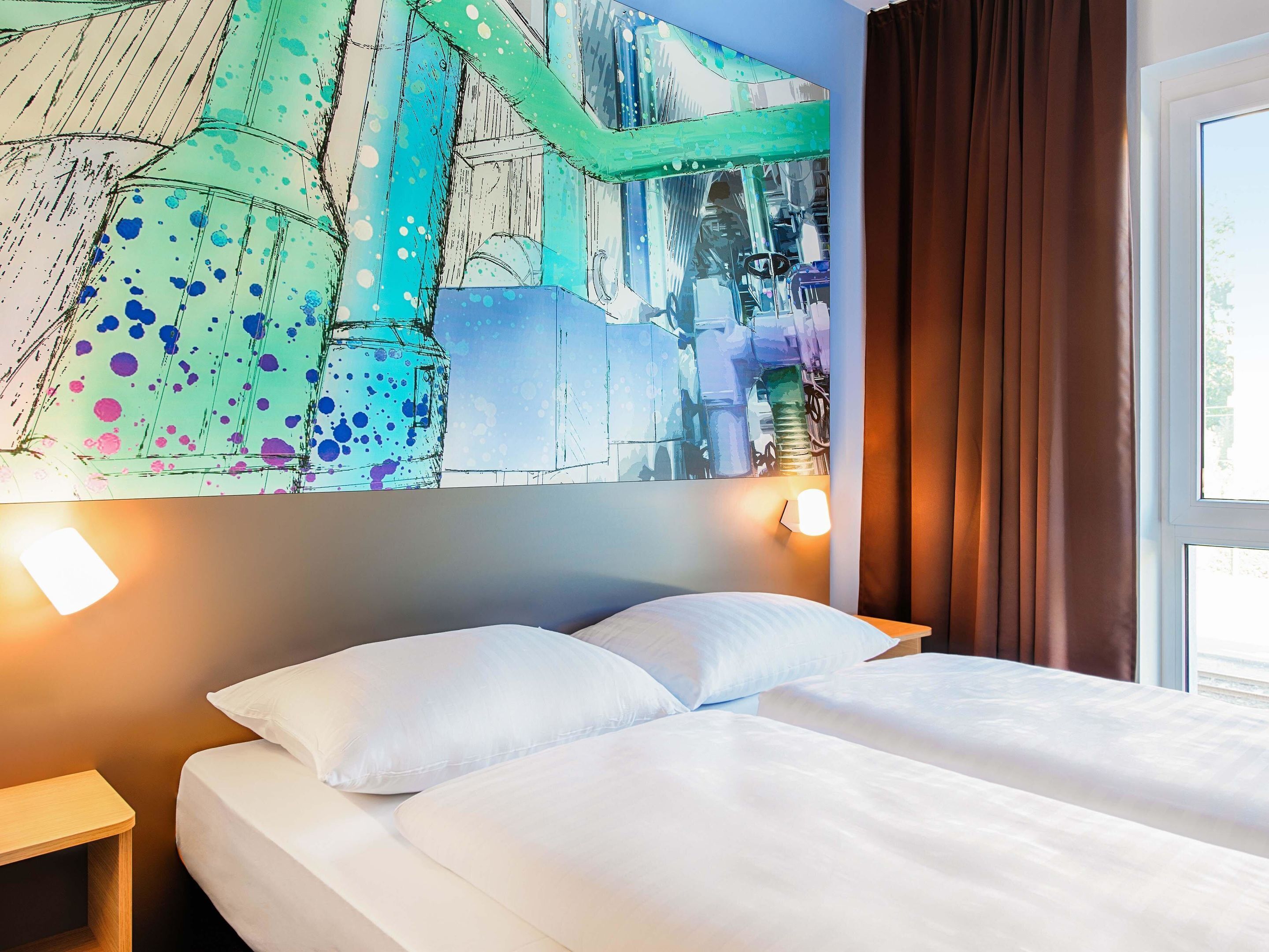 Quarto de hotel com uma cama, almofadas brancas, uma mesa de cabeceira, parede com pintura abstrata colorida e uma janela com cortinas.
