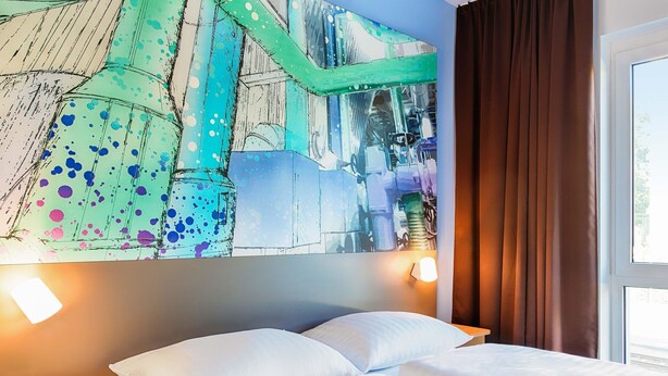 A hotel room with a bed, white pillows, a nightstand, a wall with a colorful abstract painting, and a window with curtains. - B&B Hotel Chemnitz (Chemnitz)