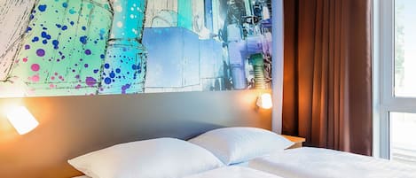 A hotel room with a bed, white pillows, a nightstand, a wall with a colorful abstract painting, and a window with curtains.