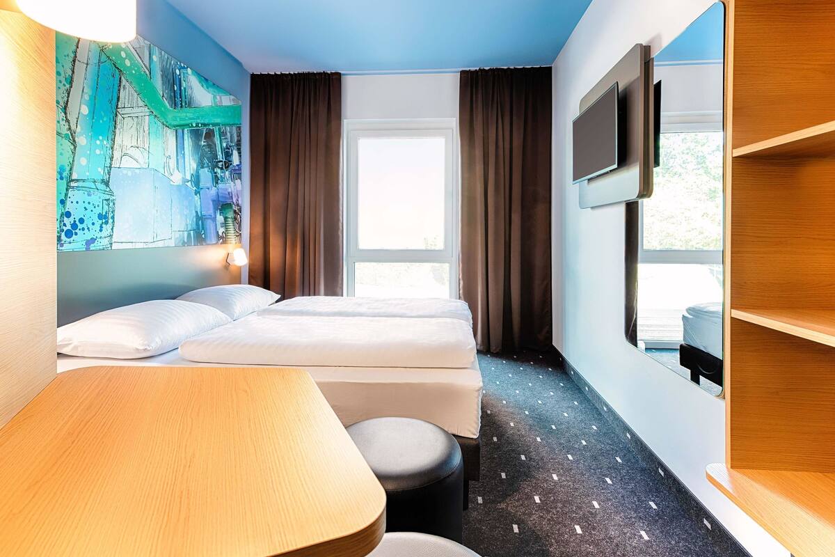 Una habitación de hotel con dos camas, un escritorio, un televisor y un ventanal grande.