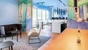 A modern interior with a large wooden table, a white chair, and a sofa. There is a glass partition with a colorful abstract mural. - B&B Hotel Chemnitz (Chemnitz)