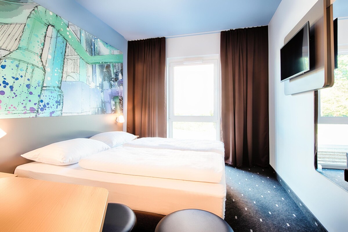 A hotel room with a bed, a desk, a chair, a TV, and a window with curtains.