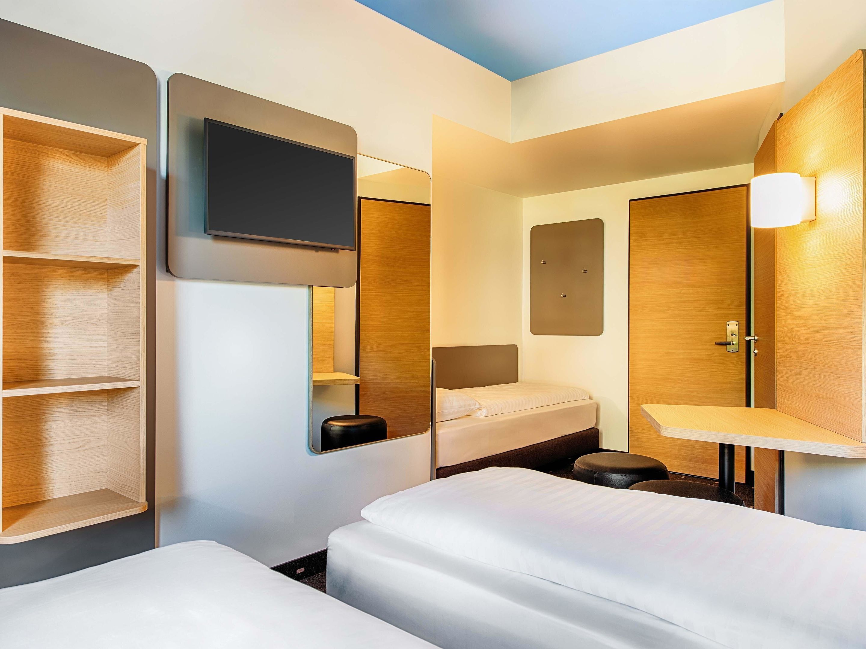 A modern hotel room with two beds, a TV, and built-in shelving.
