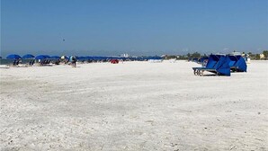 Ein Strand mit blauen Sonnenschirmen und Liegestühlen, ein klarer Himmel und ein Blick auf Gebäude in der Ferne.