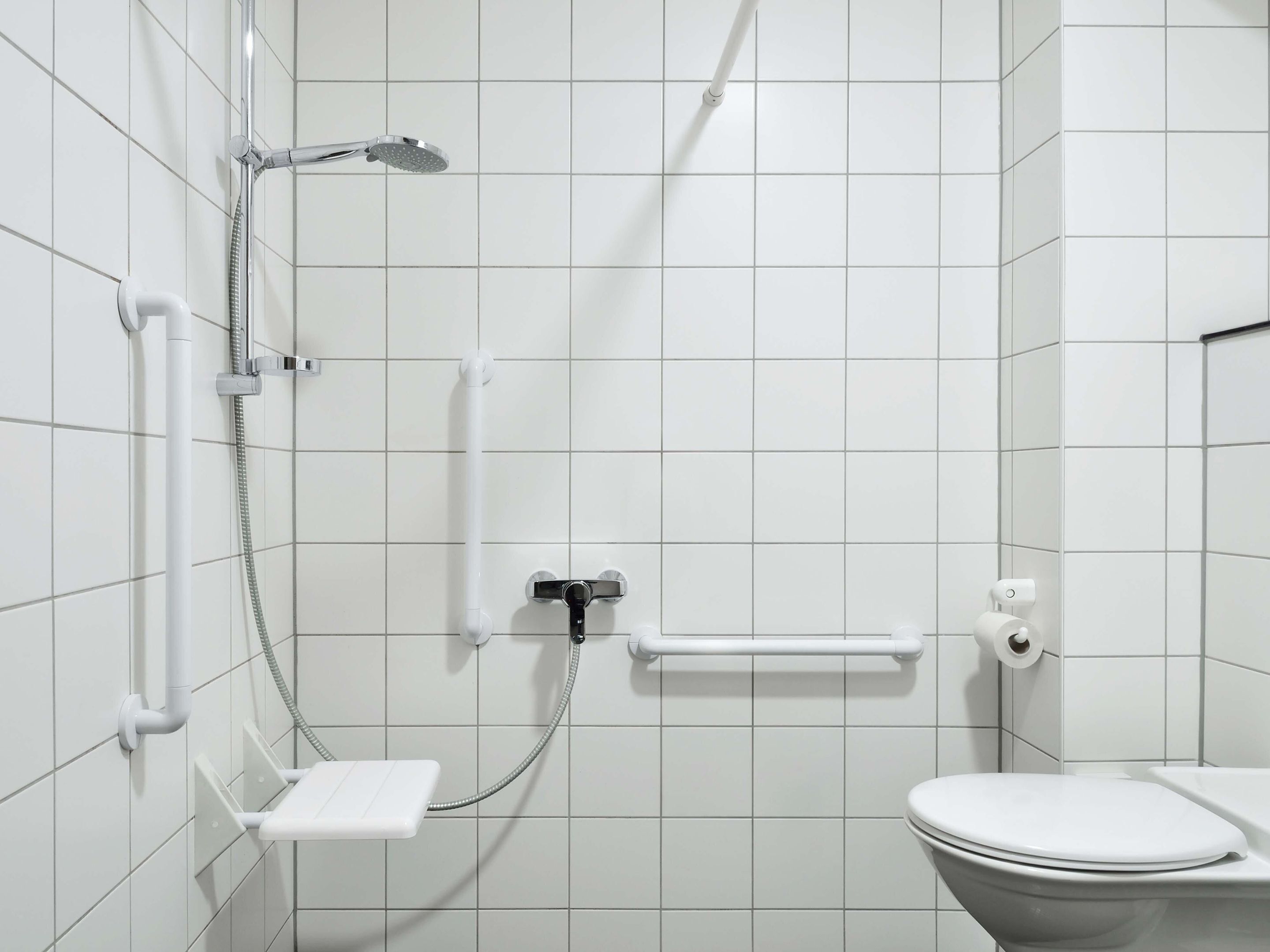 Ein modernes Badezimmer mit weißen Fliesen, einer Dusche inklusive Handbrause, einer Toilette und einer Haltestange.