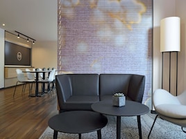 Ein modernes Hotelzimmer mit einer Eckcouch, zwei runden Tischen, einem weißen Sessel, einem großen abstrakten Wandbild und einem Schreibtisch mit Stühlen.