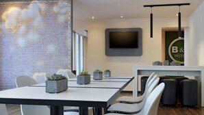 A modern hotel room with a dining area featuring a table and chairs, a flat-screen TV mounted on the wall, and a reception desk with a sign. - B&B Hotel Düsseldorf-Ratingen (Ratingen)