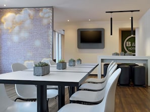 Habitación de hotel moderna con zona de comedor que incluye una mesa y sillas, un televisor de pantalla plana en la pared y un mostrador de recepción con un cartel.