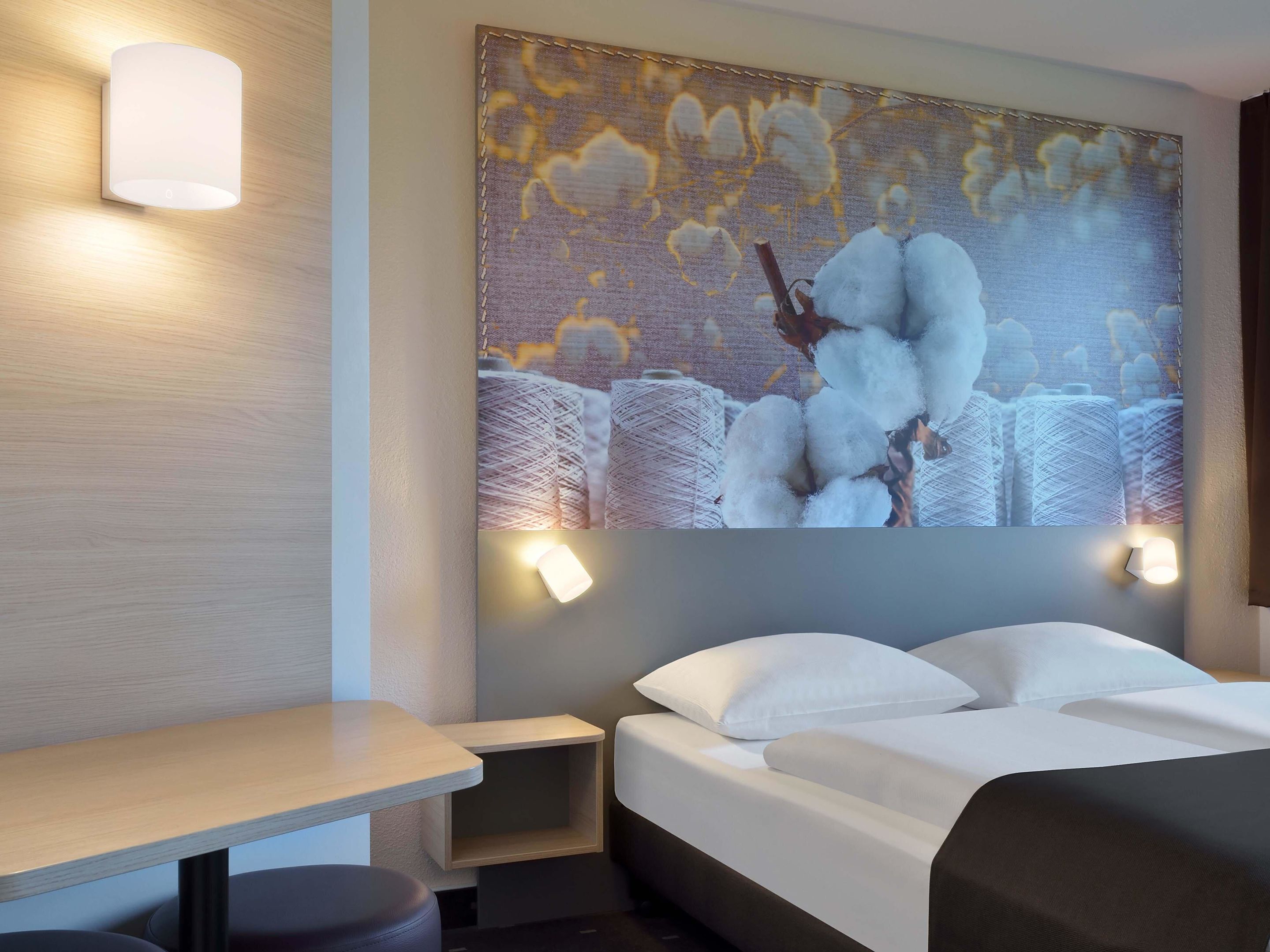 A hotel room with a bed, a nightstand, a wall-mounted lamp, and a large cotton-themed artwork.
