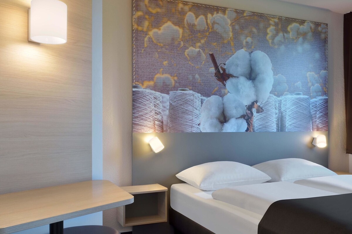 A hotel room with a bed, a nightstand, a wall-mounted lamp, and a large cotton-themed artwork.